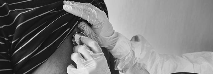 Cropped close-up snapshot of doctor in PPE protective suit examining elderly male patient at home using phonendoscope, checking chest, black and white. Coronavirus concept