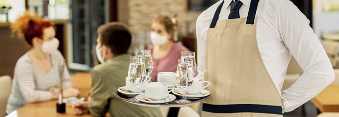 Happy waiter serving coffee to his customers while wearing protective face mask in a cafe due to coronavirus epidemic.